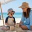 «Μητέρα και παιδί προστατευμένα με καπέλα, αντηλιακό και μπλουζάκι UV παίζουν στη σκιά μιας ομπρέλας σε ελληνική παραλία.»