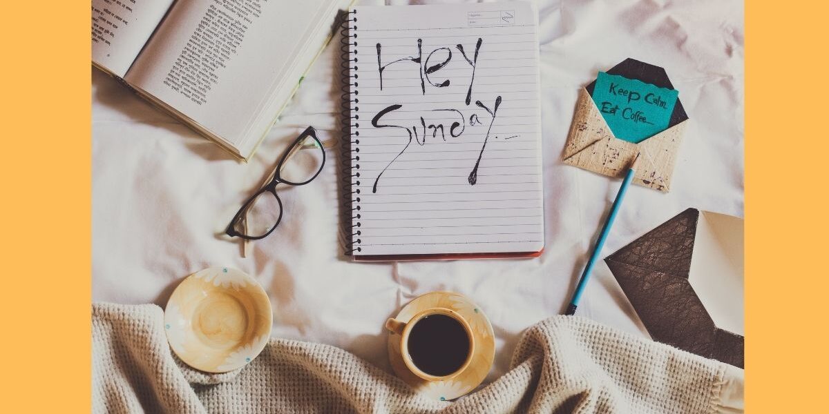 Flat lay φωτογραφία σε λευκά σεντόνια με έναν καφέ, ένα βιβλίο, γυαλιά και ένα τετράδιο που γράφει "Hey Sunday".