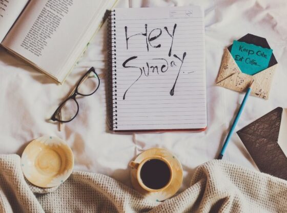 Flat lay φωτογραφία σε λευκά σεντόνια με έναν καφέ, ένα βιβλίο, γυαλιά και ένα τετράδιο που γράφει "Hey Sunday".