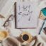 Flat lay φωτογραφία σε λευκά σεντόνια με έναν καφέ, ένα βιβλίο, γυαλιά και ένα τετράδιο που γράφει "Hey Sunday".